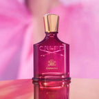 Creed Carminia perfume bottle on a reflective surface with a blurred pink background